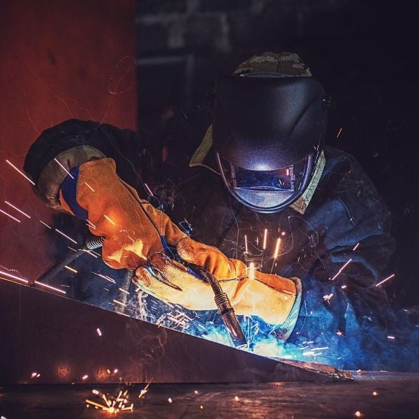 A welder welding with proper safety gear. The image is dark with blue and red hues from the sparks.